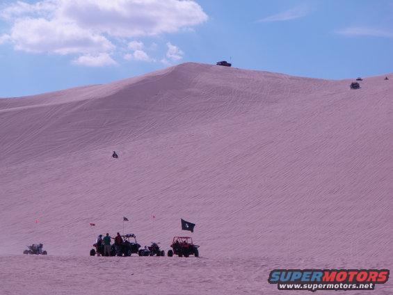 Several off-road vehicles and people gathered on a large sand dune under a blue sky with clouds. Some vehicles are driving on the dune