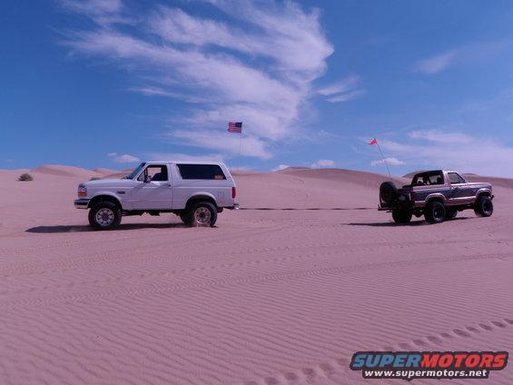 Two Ford Bronco SUVs driving on sand dunes under a blue sky with clouds. One white Bronco is in the foreground, and a darker Bronco is behind it