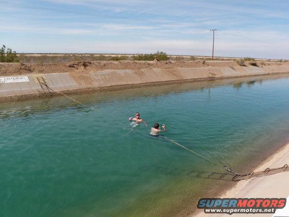 Two people swim in a wide, concrete canal under a bright blue sky. Desert landscape borders the canal