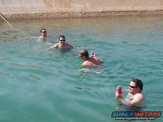 Four men swim in a canal, two holding cans of soda