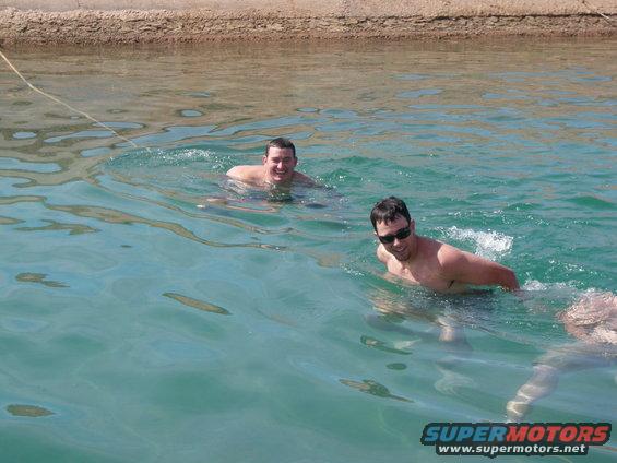 Two men swim in clear blue water near a sandy shore, with a 'SUPERMOTORS' watermark in the lower right
