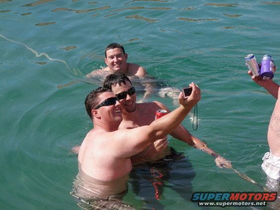 Three men in the water, two taking a selfie with a camera, one holding cans