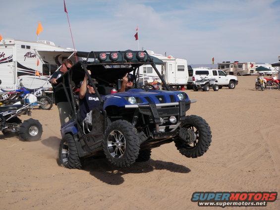 Blue side-by-side off-road vehicle with two people, one giving a middle finger. Other ATVs and RVs in background