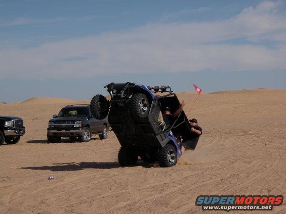 Side-by-side ATV flipping onto its side in the sand with two people trying to hold it up. Another SUV and truck are parked nearby