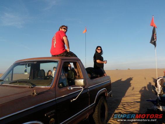 Brown 1980s Ford Bronco with people on roof, desert scene with flags and another vehicle