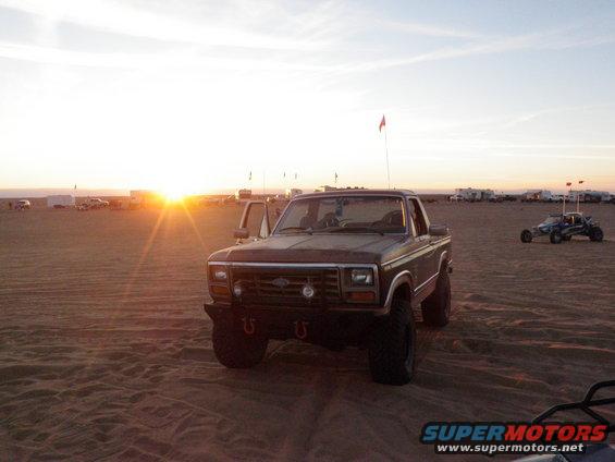 Brown Ford pickup truck parked on sand dunes at sunset, with a dune buggy in the background