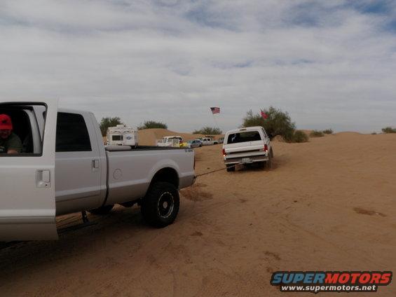 White Ford Bronco being towed by a white pickup truck in a sandy desert landscape. American flag visible in background