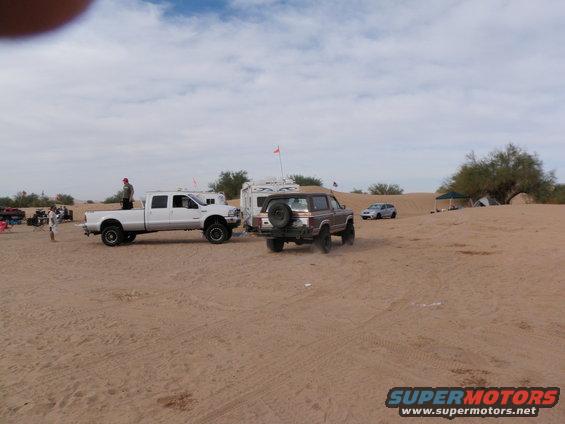 Brown Ford Bronco with spare tire on back, parked next to white pickup truck and trailer in sandy desert