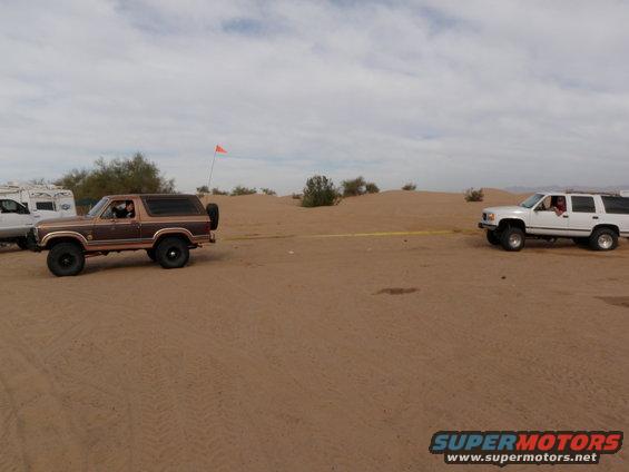 Brown Ford Bronco with a spare tire on the back, being towed by a white SUV in a sandy desert