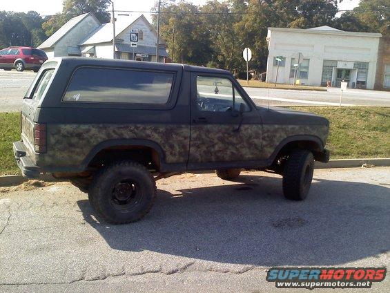 Side view of a lifted Ford Bronco with camouflage paint. It's parked on asphalt near some buildings