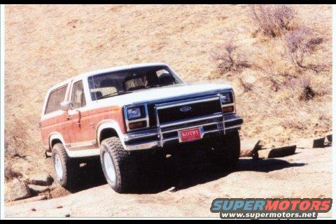 1984 Ford Bronco driving on a dirt road with a red license plate that says KOTBY