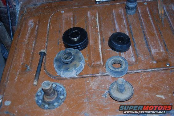 Several rubber and metal suspension parts laid out on a brown, ribbed surface. Includes a long bolt, a spring-loaded mount, and various bushings