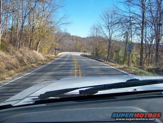 View from inside a vehicle looking out at a two-lane road winding through autumn trees under a clear blue sky