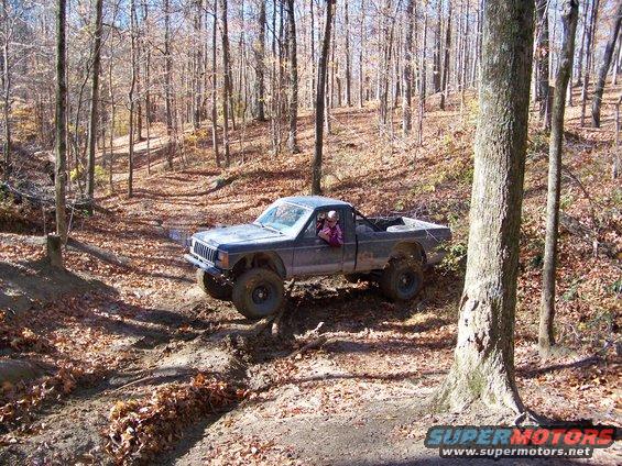 Jeep Comanche pickup truck with large tires driving on a dirt trail in a wooded area