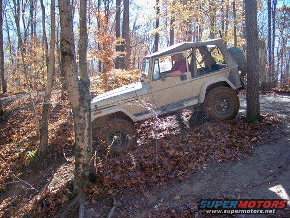 Tan Jeep Wrangler on wooded trail, driver visible, soft top up, tires muddy