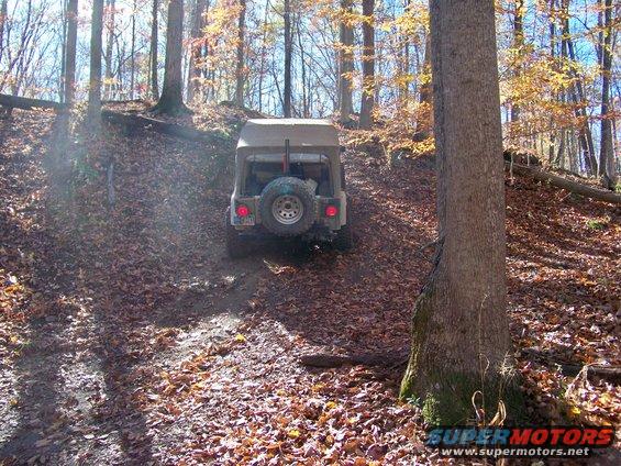 Jeep CJ-7 with canvas top driving uphill on a dirt trail covered in autumn leaves