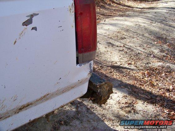 Rear corner of a white pickup truck with rust spots and mud. Exhaust pipe is resting on a log
