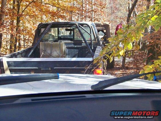 Pickup truck with roll cage and auxiliary fuel tank in wooded area. Another vehicle is partially visible behind it