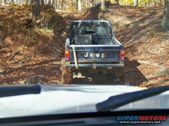 Rear view of a black Jeep pickup truck with 'Jeep' on tailgate, off-roading on a dirt path with fallen leaves. Another vehicle's windshield is in the foreground