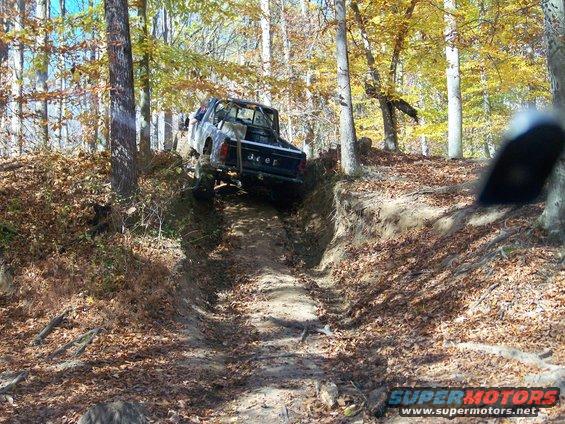 Black Jeep truck with a roll bar climbing a steep, dirt trail in a forest with autumn leaves