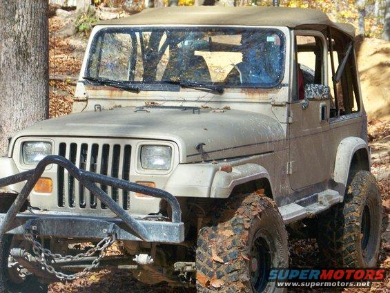 Tan Jeep Wrangler with a brush guard and large mud tires, parked on a dirt path with trees around