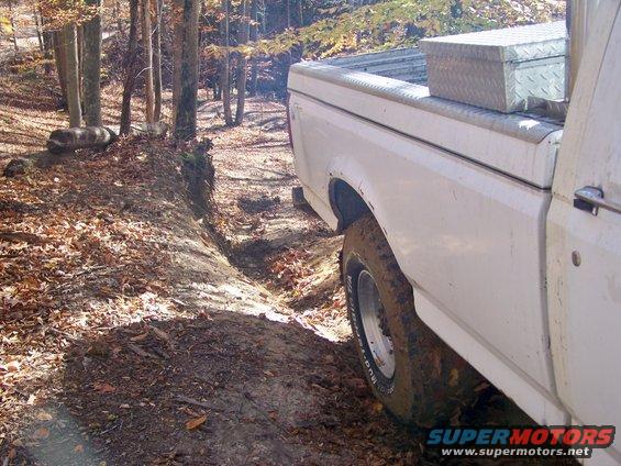 White 1987 Ford F-250 driving on a dirt path in a forest, rear passenger side visible