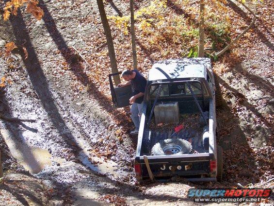 Jeep pickup truck with roll cage stuck in muddy terrain, spare tire in bed