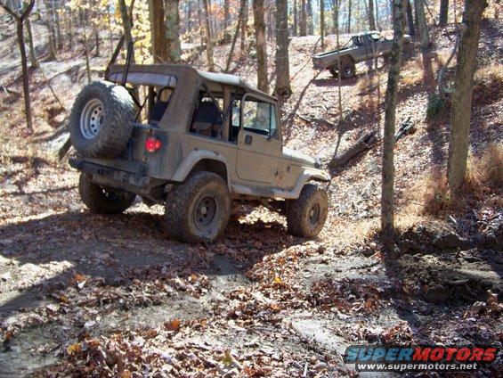 Tan Jeep Wrangler with spare tire on rear, navigating a wooded trail with another truck behind