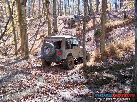 Tan Jeep Wrangler and a pickup truck driving on a wooded trail. The Jeep is in the foreground, facing away from the camera