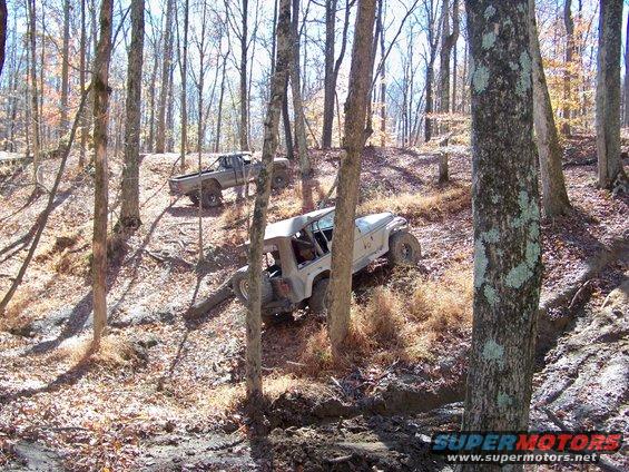 Two off-road vehicles navigate a muddy trail through a forest with bare trees
