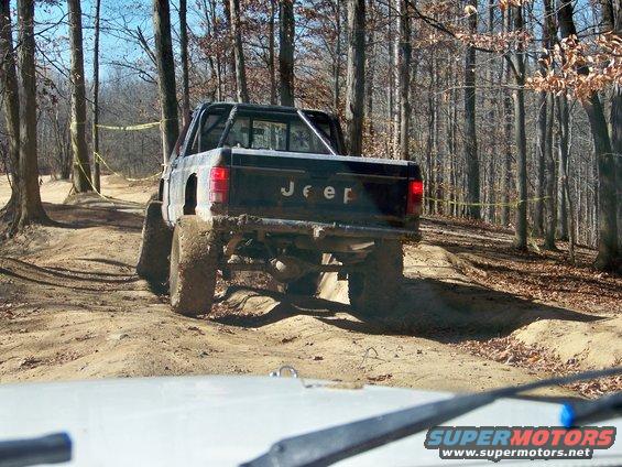 Black Jeep pickup truck with large tires on a dirt trail, rear view