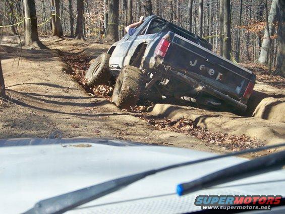 Jeep pickup truck stuck on muddy trail in woods; another vehicle's windshield wipers in foreground