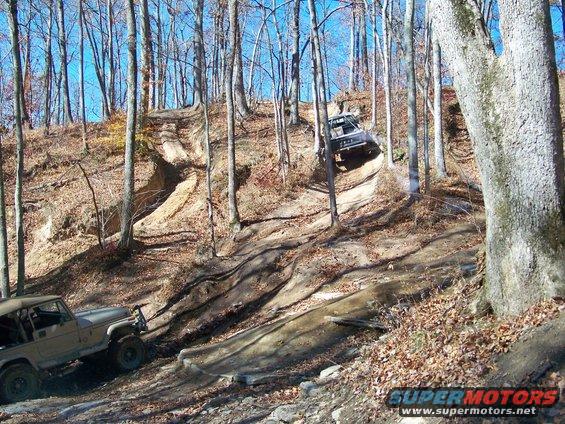 Two off-road vehicles navigate a steep, wooded trail with fallen leaves