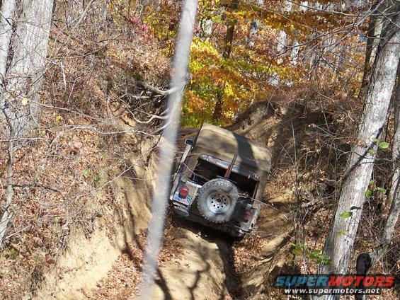 Tan Jeep Wrangler on a dirt trail, spare tire mounted on rear, autumn foliage