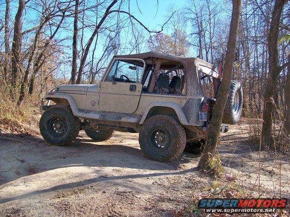 Tan Jeep Wrangler with a soft top, spare tire mounted on the rear, on a dirt trail in a wooded area