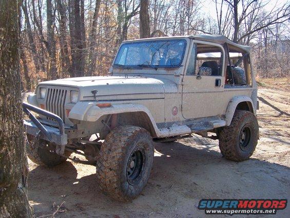 Tan Jeep Wrangler with aftermarket bumper and large tires, parked on dirt road