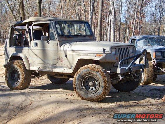Tan Jeep Wrangler YJ with off-road tires parked on a dirt trail next to a dark SUV