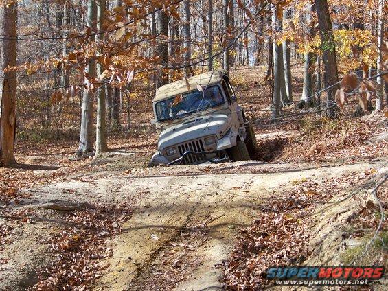 Tan Jeep Wrangler stuck in a ditch on a dirt path, surrounded by trees with fall foliage