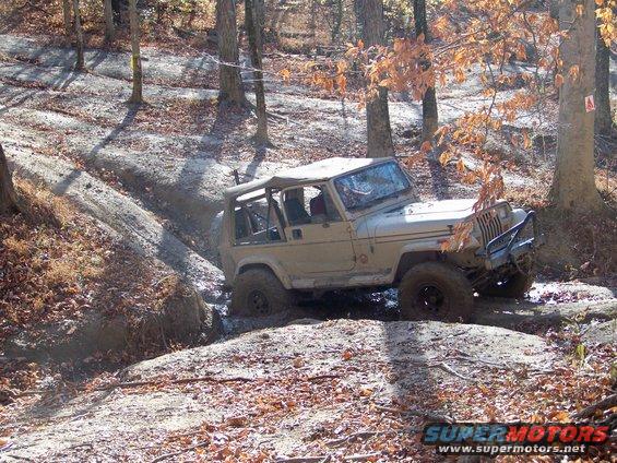 Tan Jeep Wrangler navigating a muddy trail in a wooded area