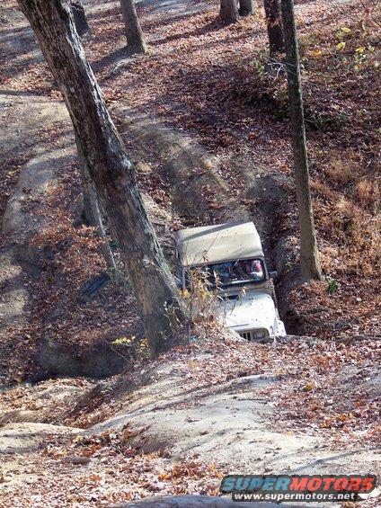 White Jeep Wrangler with a tan soft top navigating a steep, wooded trail with fallen leaves