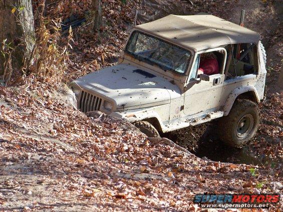 Tan Jeep Wrangler off-roading on a dirt path covered in leaves, with the front driver's side wheel stuck in mud