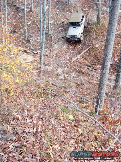 Jeep Wrangler driving down a steep, wooded trail in autumn. The vehicle is tan with a white top