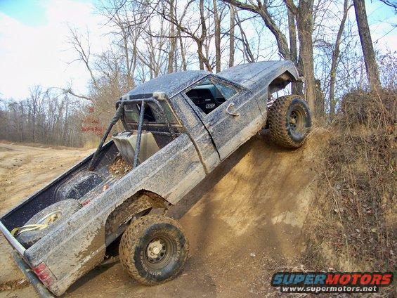 Mud-covered 1986 Jeep Comanche pickup truck with a roll cage, climbing a dirt hill