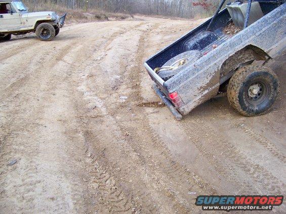Muddy pickup truck bed and rear tire on dirt track; another vehicle visible in background