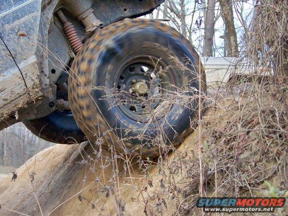 Off-road vehicle tire climbing a dirt mound with another vehicle visible in the background