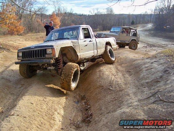 Muddy Jeep Comanche pickup truck driving off-road with a Jeep Wrangler behind it. A man waves from the side of the trail