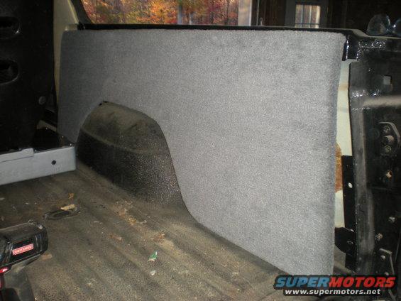 Interior rear quarter panel and wheel well covered in grey carpet. Floor shows dirt and a power tool