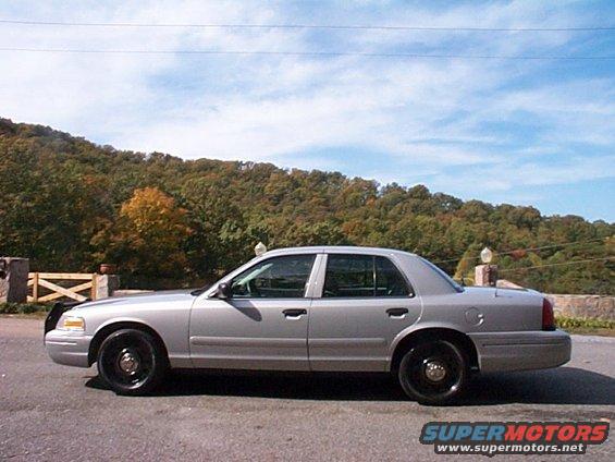 Side view of a silver 2004 Ford Crown Victoria Police Interceptor parked outdoors with trees and a hill in the background
