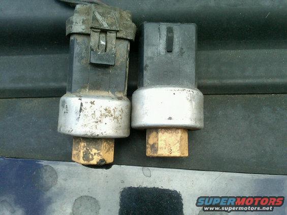 Two automotive pressure sensors, one with a black connector and silver base, the other with a black connector and silver base, resting on a dark surface