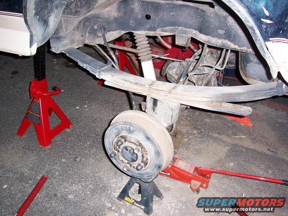 vehicle rear axle on jack stands with leaf springs and shock visible, person working underneath